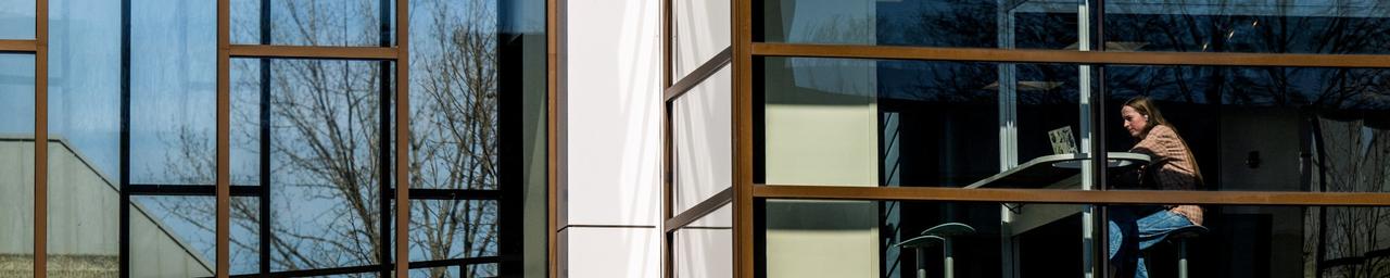 The outside of Au Sable looking at the glass windows and a student sitting inside working on their laptop